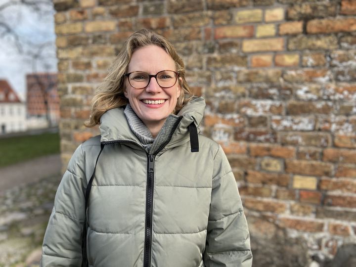 Eine Frau mit Brille steht vor einer Hauswand und blickt freundlich in die Kamera.