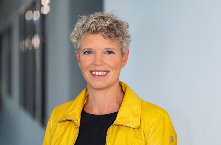 Eine Frau mit kurzen Haaren, gelber Jacke und schwarzem Oberteil schaut freundllich in die Kamera.