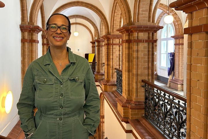 Eine Frau mit grüner Bluse steht in einem historischen Treppenhaus und schaut in die Kamera. 
