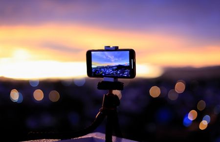 eine auf einem Stativ aufgestellte Kamera ins Tal gerichtet, wo man in der Ferne Licher eines Ortes und einen Sonnenuntergang sieht - Videofunktion