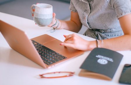 Ausschnitt einer jungen Frau, die vor dem Laptop sitzt mit Kaffeetasse und Block