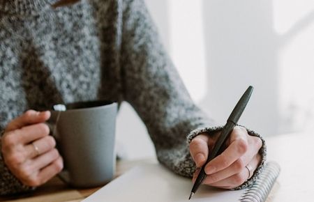 Ausschnitt eines Oberkörpers einer Frau, die mit einer Hand eine Teetasse hält, in der anderen einen Stift. Vor ihr liegt ein aufgeschlagener Block