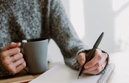 Ausschnitt eines Oberkörpers einer Frau, die mit einer Hand eine Teetasse hält, in der anderen einen Stift. Vor ihr liegt ein aufgeschlagener Block