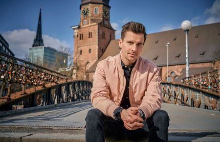 Ein Mann mit rosefarbener Jacke sitzt im Freien, im Hintergrund eine Brücke, blauer Himmel und Gebäude.