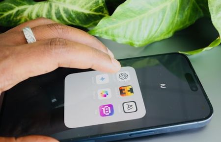 Ausschnitt einer beringten Hand tippt auf ein Handyscreen mit KI-Symbolen vor planzlichem Hintergrund