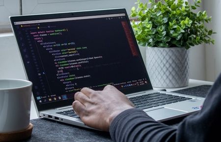 Ein aufgeklappter Laptop auf Schreibtisch vor Fenster, an dem eine Hand die Tastatur bedient und promptet