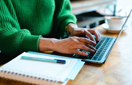 Ausschnitt einer Frau im grünen Strickpulli, die ihre Hände am Laptop hat, daneben ein Notizblock mit Stift