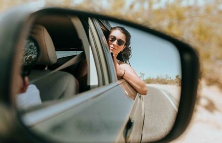 Im fahrenden Auto sieht man im Rückspiegel eine Frau, die mit Sonnenbrille rausschaut und den Ellenbogen aus dem Fenster hängt - Urlaubsvibes