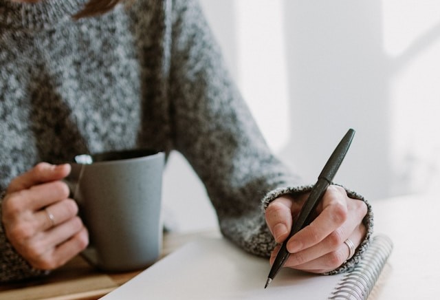 Kelly Sikkema/ unsplash Ausschnitt eines Oberkörpers einer Frau, die mit einer Hand eine Teetasse hält, in der anderen einen Stift. Vor ihr liegt ein aufgeschlagener Block